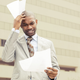 A man in a business suit in front of a building rubs his head in befuddlement at papers he is reading.